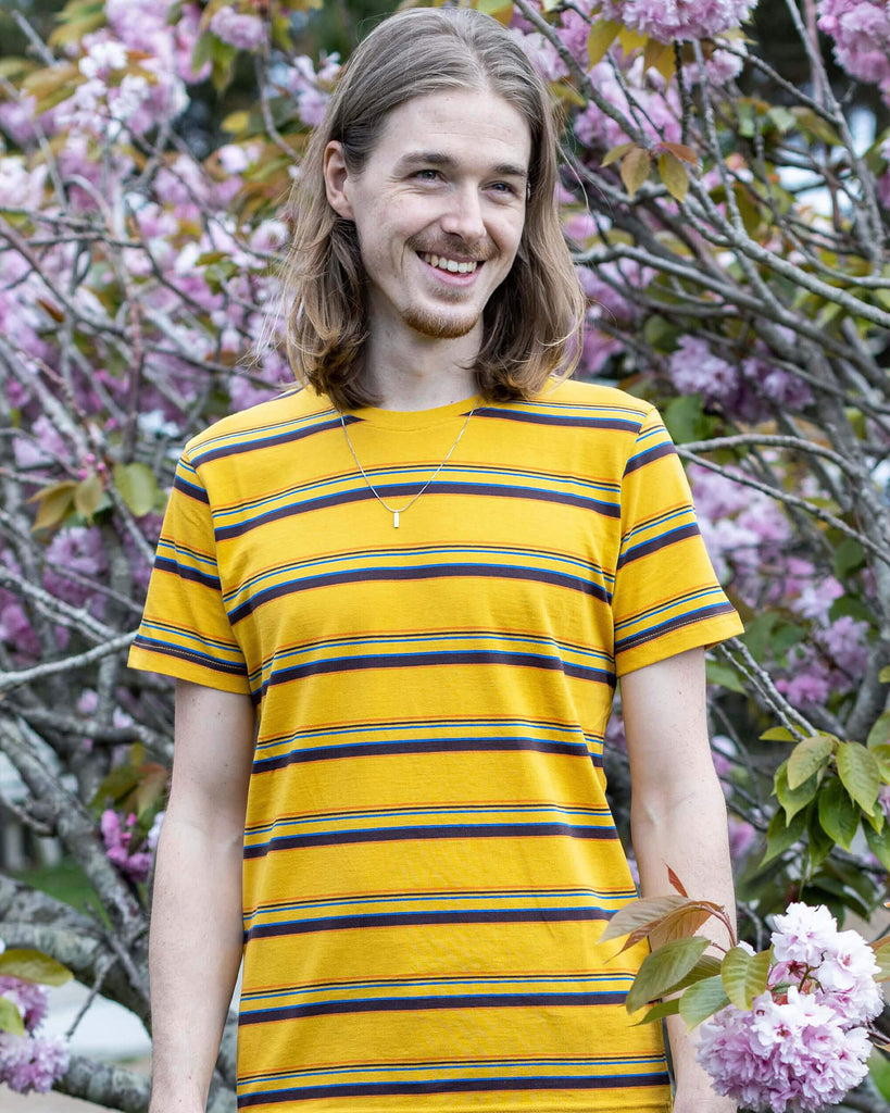 Jack is stood in front of a blossom tree wearing the gold sunset stripe retro tshirt with tobacco corduroy flares. He has mid length blonde hair and is smiling facing towards the camera with both hands resting by his sides. Photo is cropped from the knees up.