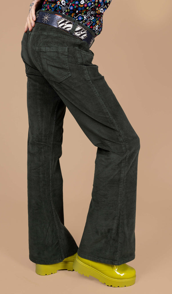 Close up of James wearing the forest green corduroy bell bottom flares with the boogie bones shirt and yellow boots. He is facing right with one leg bent forward to highlight the flare shape of the trousers. The photo is cropped from the hips down.
