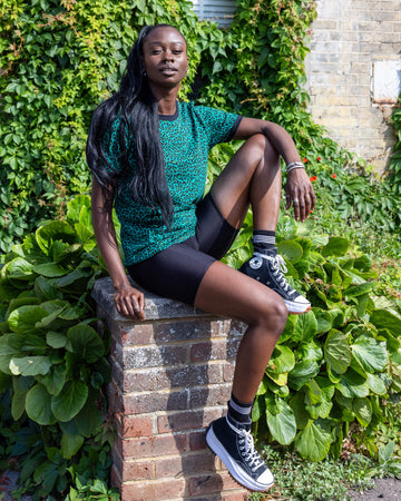 Yolande is wearing Teal Leopard Print T Shirt with black trousers. She is stood outside in Hove on a sunny day smiling at the camera and holding onto the bottom of the t shirt.