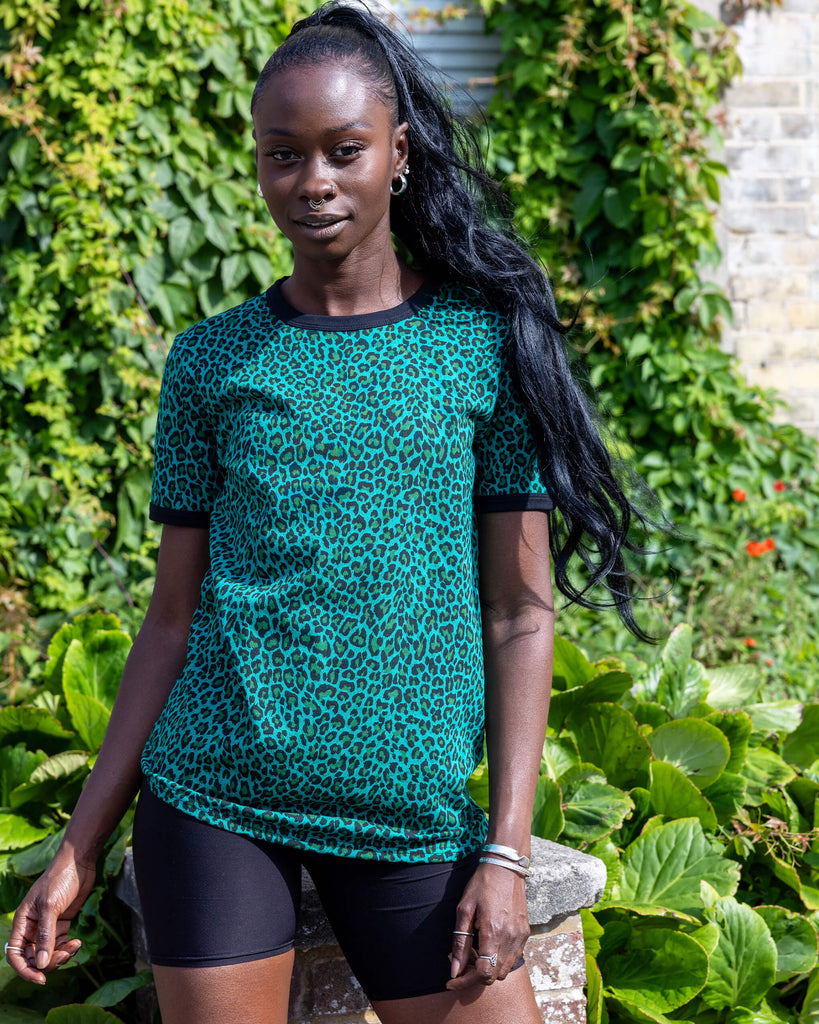 Yolande is wearing Teal Leopard Print T Shirt with black trousers. She is stood outside in Hove on a sunny day smiling at the camera and holding onto the bottom of the t shirt.
