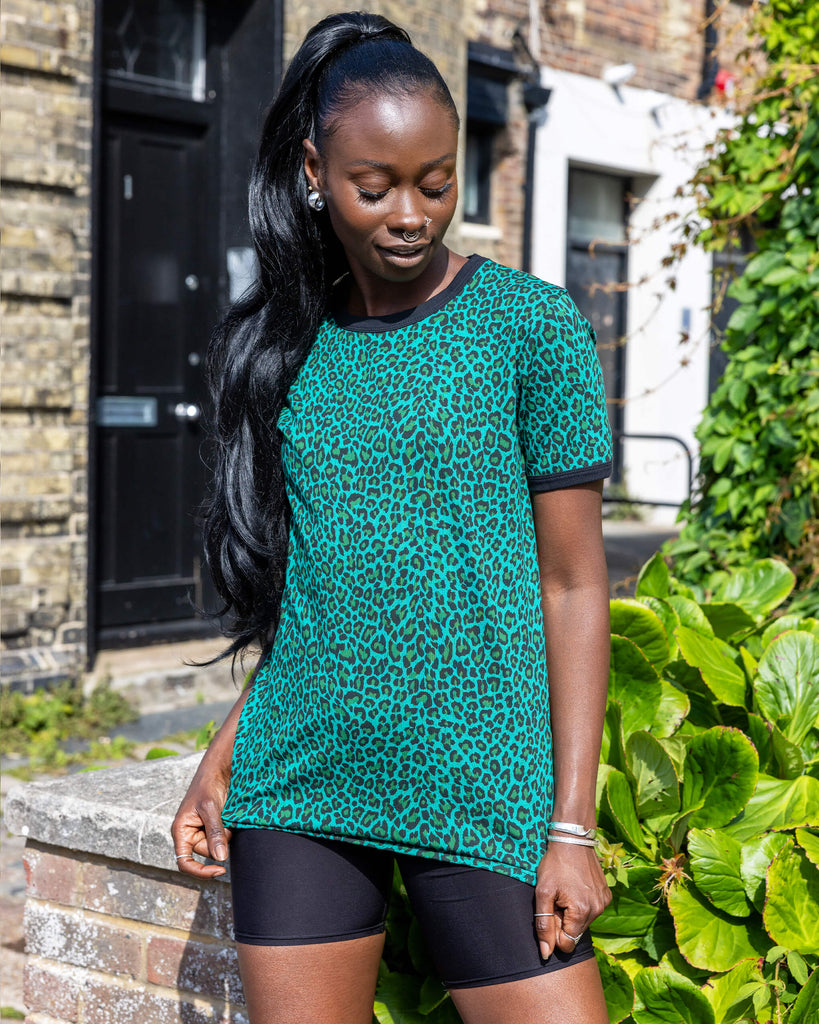 Yolande is wearing Teal Leopard Print T Shirt with black trousers. She is stood outside in Hove on a sunny day smiling at the camera and holding onto the bottom of the t shirt.