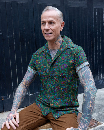 white male heavily tattooed model wearing Forest Green Paisley Short Sleeve Rayon Shirt paired with brown corduroy trousers. The shirt is a forest green colour with an all over paisley print in various muted rainbow colours. The model is sat on a wooden stood outside in Hove with a black door behind him. 