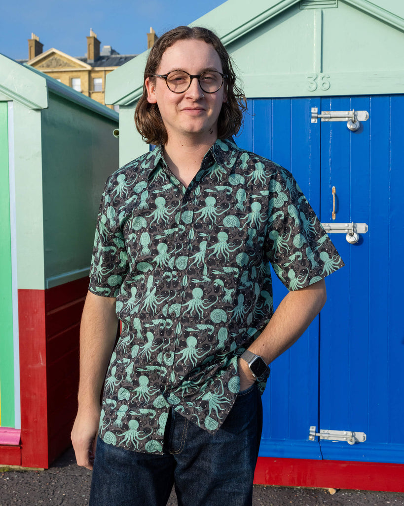 Michael is stood outside in Hove in front of beach huts wearing Run & Fly Green Seas short sleeve shirt with dark blue jeans. The shirt features a green octopus, squid, and nautilus print. Michael is posing toward the camera with one hand in his pocket and other down by his side. 