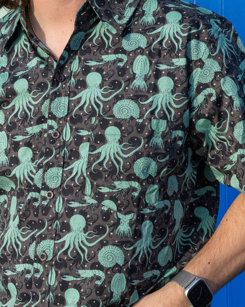 Close up of model wearing Run & Fly Green Seas short sleeve shirt with dark blue jeans. The shirt features a green octopus, squid, and nautilus print. Close up shows the front pocket on the shirt 