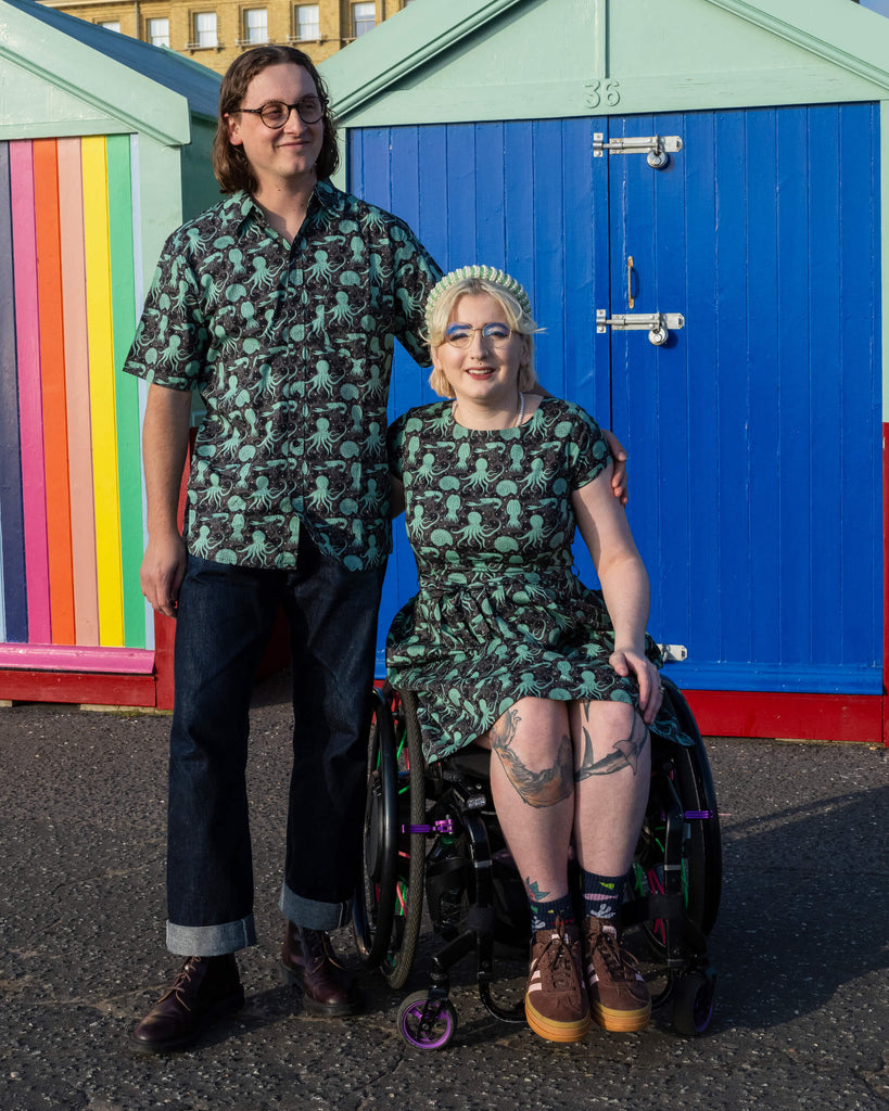 Michael are Eliza are posing in Hove of front of beach huts. Michael is wearing Run & Fly Green Seas sleeve shirt with dark blue jeans whilst Eliza is wearing Run & Fly Green seas Tea Dress with brown trainers. The dress and shirt features a green octopus, squid, and nautilus print. Eliza is sat in their wheelchair smiling at the camera while Michael stands next to them. They have their arms round each other.