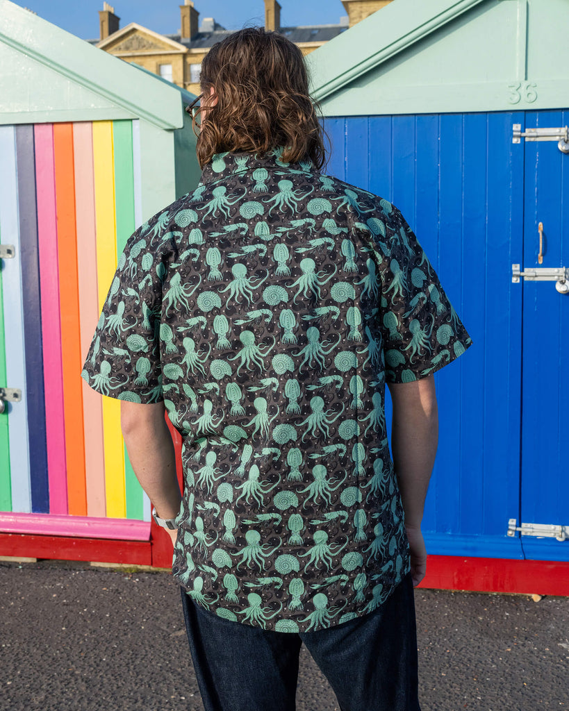 Michael is stood outside in Hove in front of beach huts wearing Run & Fly Green Seas short sleeve shirt with dark blue jeans. The shirt features a green octopus, squid, and nautilus print. He is stood with his back to the camera.