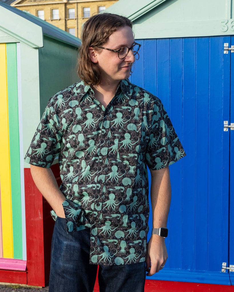 Michael is stood outside in Hove in front of beach huts wearing Run & Fly Green Seas short sleeve shirt with dark blue jeans. The shirt features a green octopus, squid, and nautilus print. They are posing looking off to one side with one hand in their pocket and other down by their side.