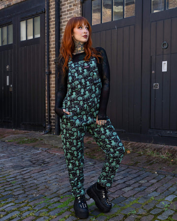 A model with tattoos and red hair stands in a relaxed pose, wearing Run & Fly black dungarees featuring a fun mint green sea creatures print. She pairs the dungarees with a black sheer top and platform boots. Run & Fly, a small ethical slow fashion brand, is known for its alternative, eco-conscious designs.