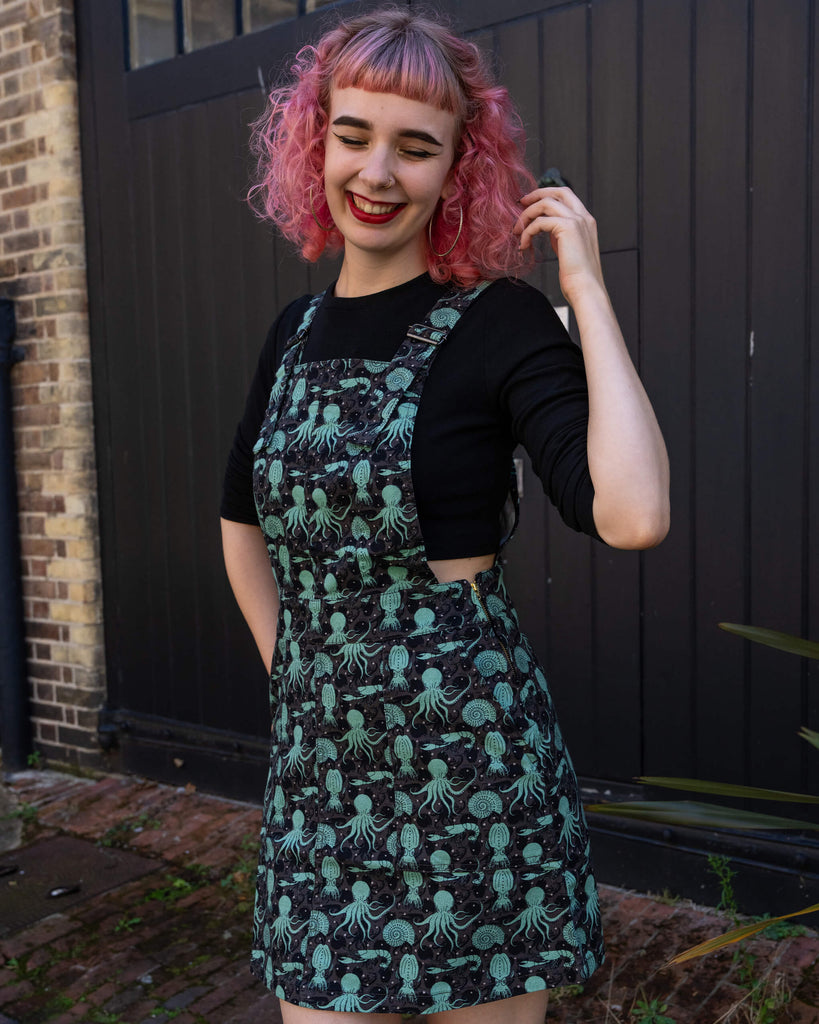Woman posing in the Run & Fly green sea creatures pinafore dress, smiling with hand in her pink curly hair. Black background contrasts with her red socks and platform shoes, highlighting the playful sea creature design.