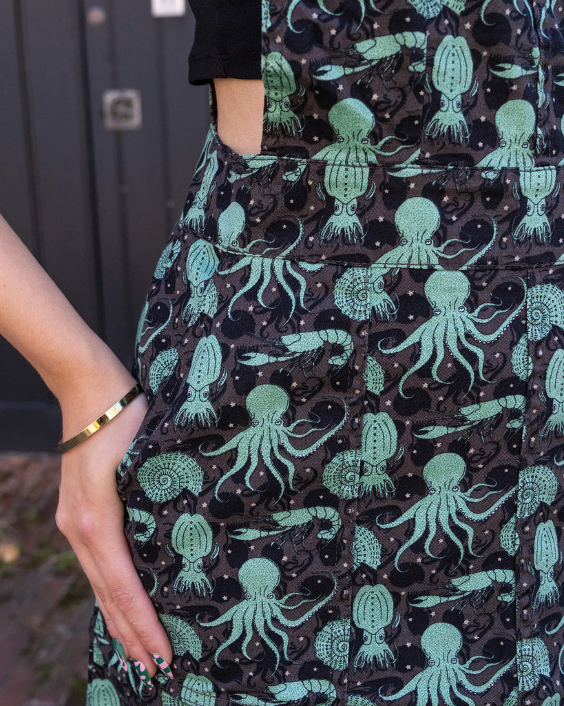 Close-up of the green sea creatures print on Run & Fly’s pinafore dress. The black corduroy fabric is adorned with whimsical octopus, squid, and nautilus designs, part of Run & Fly's ethical and sustainable fashion collection.