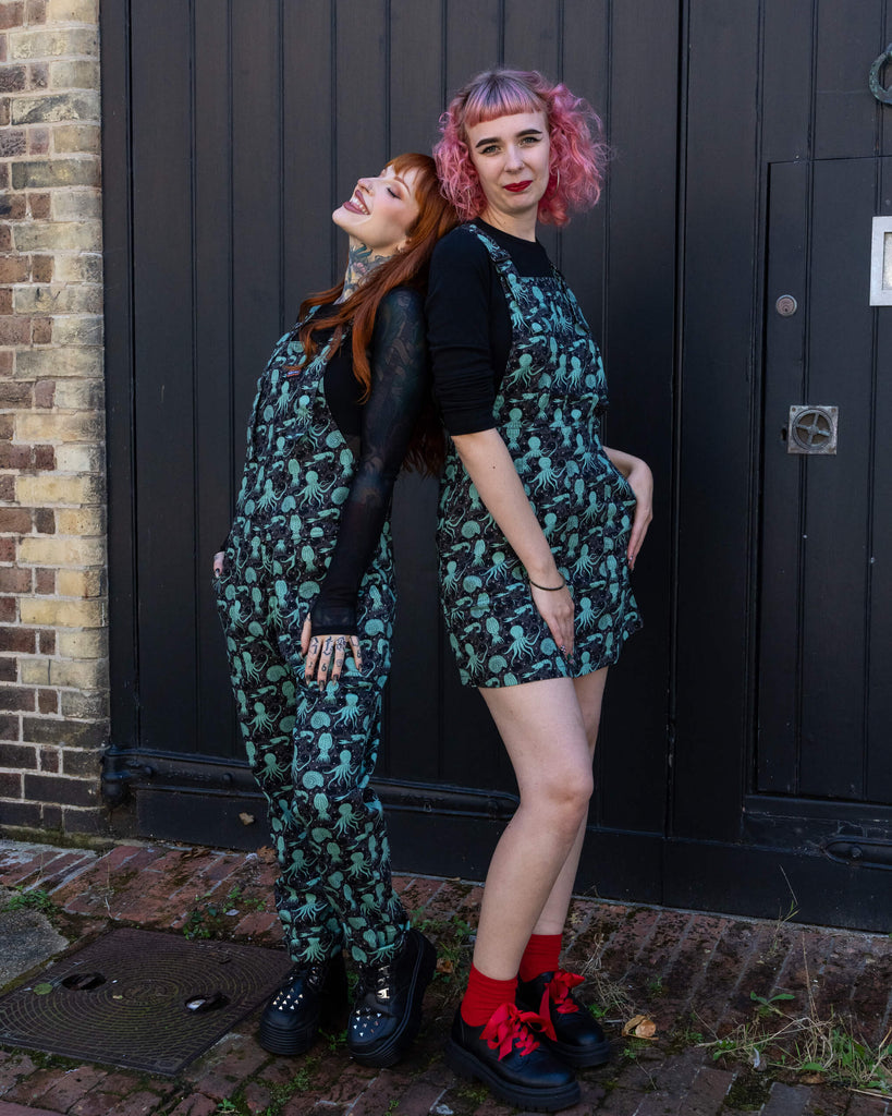 Two women pose playfully in matching Run & Fly green sea creatures outfits. One wears dungarees, the other a pinafore dress, both showing off the octopus and squid pattern.