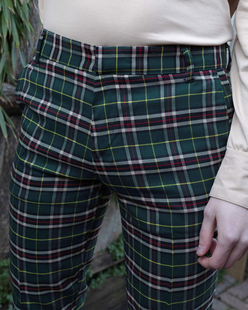 Close-up of Run & Fly Retro Mod Stretch Plaid Green Tartan Slim Skinny Fitting Trousers, featuring a classic green, red, and yellow tartan pattern. The trousers are styled with a cream long-sleeved top. Jack, the model, goes by he/him pronouns. Run & Fly is a small ethical slow fashion brand known for retro-inspired and sustainable designs.