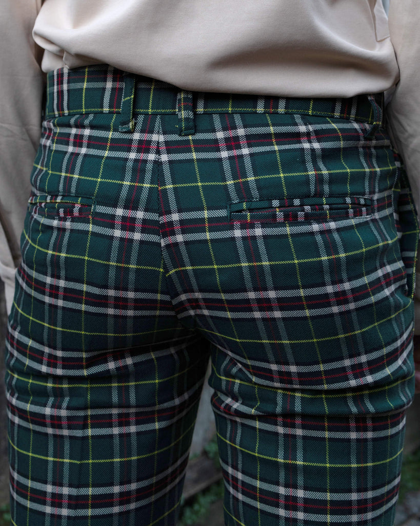Back view of the Run & Fly Retro Mod Stretch Plaid Green Tartan Skinny Fitting Trousers, showcasing the green, red, and yellow plaid pattern. The model, Jack, pairs the trousers with a cream shirt. Run & Fly offers ethical, slow fashion designs with a focus on retro style.