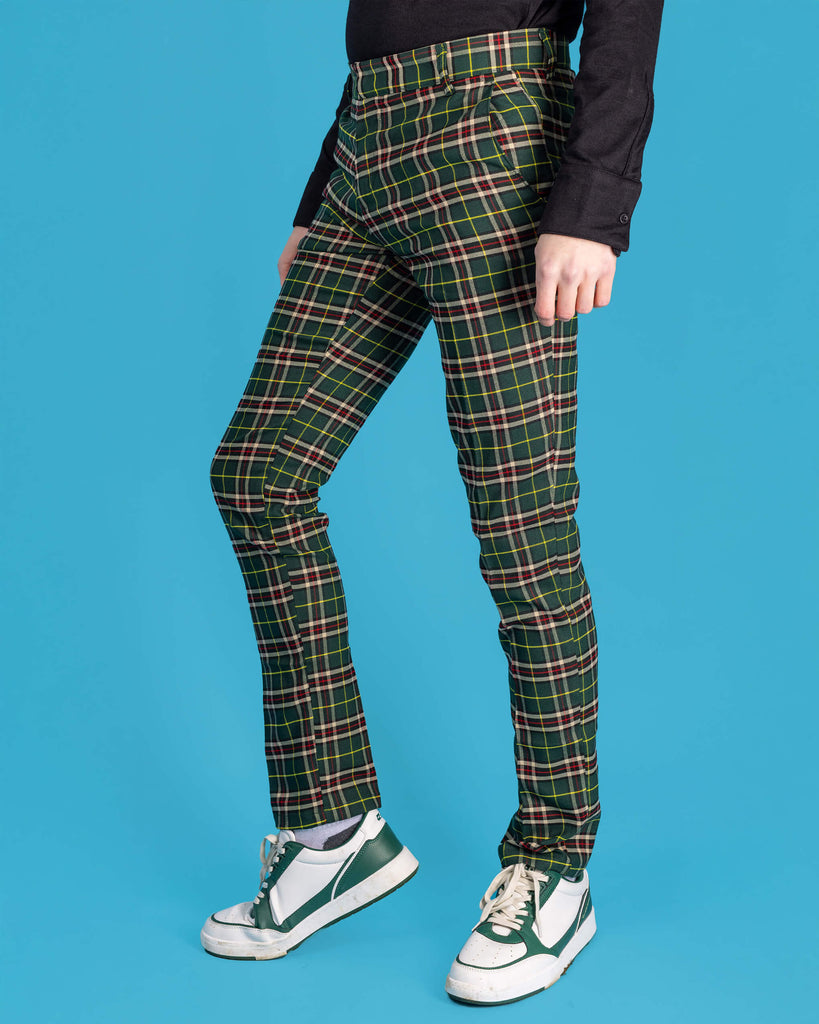 Jack is stood in front of a blue studio background wearing the retro mod stretch plaid green tartan slim skinny fitting trousers with a long sleeve black shirt and white trainers. They are facing to the left with one leg bent forward to highlight the trouser shape. Photo is cropped from the waist down.