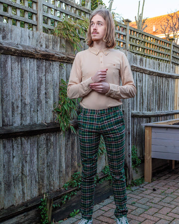 Jack is stood in a garden wearing the retro mod stretch plaid green tartan slim skinny fitting trousers with a sand coloured long sleeve shirt with white trainers. They are facing the camera posing doing up one of the shirt cuff buttons and looking off to the left.