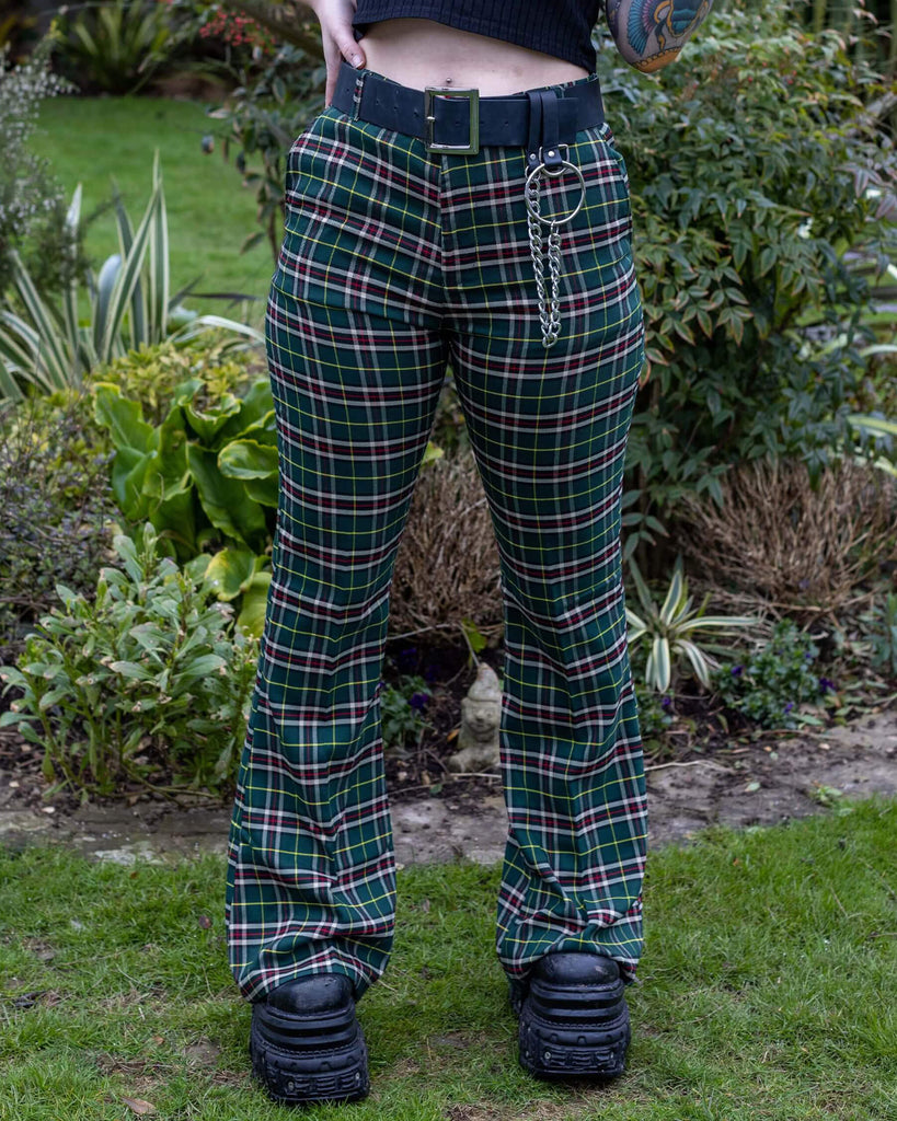 Florence is stood outside in a garden in Hove wearing Green Tartan Plaid Bell Bottom Slacks paired with a black belt with chain, and chunky black boots. She is facing the camera and posing with one hand on her hip. Photo is cropped at the waist