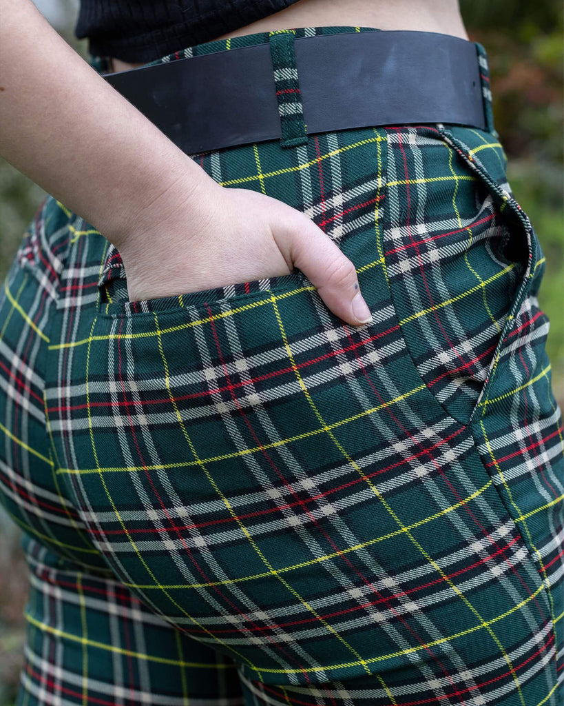 Close up of Florence stood outside in a garden in Hove wearing Green Tartan Plaid Bell Bottom Slacks paired with a black belt with chain, and chunky black boots. She is facing away from the camera and posing with one hand in the back pocket. 