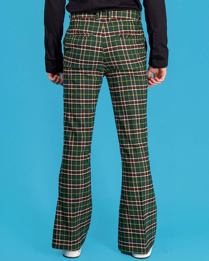 Jack is stood in front of a blue studio background wearing the green tartan plaid bell bottom slacks with a long sleeve black tshirt and white trainers. He is facing away from the camera with both hands resting by his sides. Photo is cropped from the waist down.