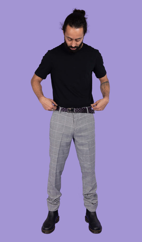 Richard, a hispanic male model with dark hair and a beard, is stood in a photography studio in front of a lilac backdrop wearing Retro Mod Prince Of Wales Stretch Slim Skinny Fitting Trousers with a black t shirt tucked in, a checkered belt and black boots. He is facing the camera looking down with his hands pulling his t shirt down slightly. 