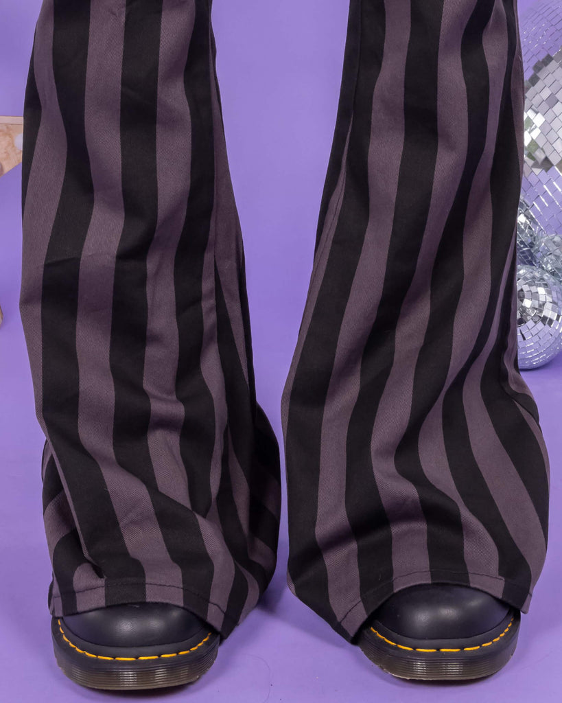 Close-up of the hem of Run & Fly grey and black vertical striped flare trousers, showing the flared silhouette that covers black Dr Martens boots. The fabric is thick and structured, with bold stripes for a statement look. Background is lilac. A unique piece from a small, ethical, slow fashion brand. Perfect for alternative style enthusiasts.