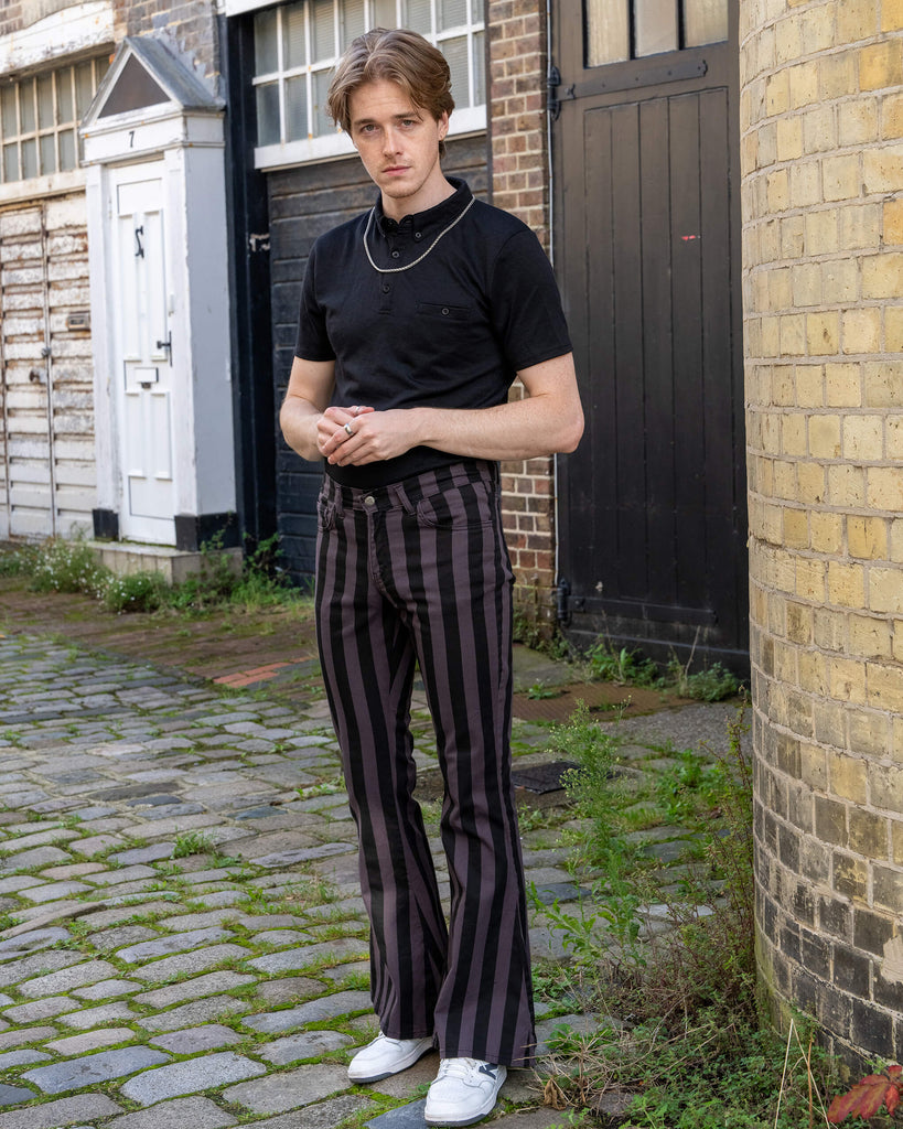 Front view of a model wearing Run & Fly grey and black vertical striped flare trousers. These high-waisted trousers feature a bold flare and vertical stripes for a retro-inspired look, paired with a simple black t-shirt and white trainers. The model stands in a cobblestone alleyway with dark wooden doors. Made ethically by a small slow fashion company in England.