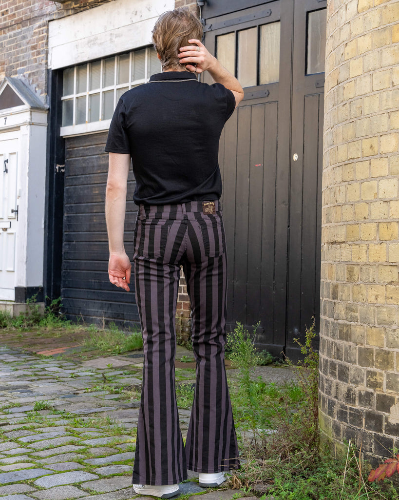 Back view of a model in Run & Fly grey and black vertical striped flare trousers. The trousers hug the waist and hips, flowing into a wide flare at the legs. The model pairs them with a casual black t-shirt and stands in a cobblestone alley with dark wooden doors. Ethical slow fashion made in England.