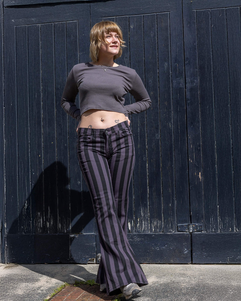 Front view of a model wearing Run & Fly grey and black vertical striped flare trousers. The high-waisted design and bold stripes flatter the figure, paired with a cropped grey long-sleeve top. The model smiles while standing against a dark wooden background. Ethically made, small slow fashion brand. Perfect for retro and alternative styles.