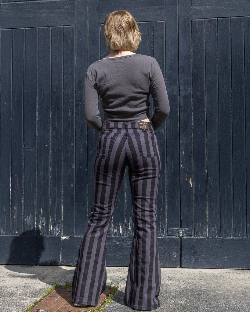 Back view of a model wearing Run & Fly grey and black vertical striped flare trousers. The high-waisted design accentuates the figure, with bold stripes creating a retro feel. The model stands confidently in front of a dark wooden panel background. Ethical slow fashion brand. Made in England. Perfect for a unique, alternative style.