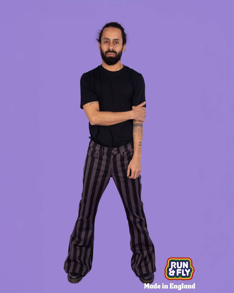 Front view of a model in Run & Fly grey and black vertical striped flare trousers. The trousers have a classic five-pocket style, high-waisted fit, and bold flare for a retro-inspired look. The model pairs them with a black t-shirt, standing confidently in front of a lilac background. Ethical slow fashion. Made in England. Alternative, retro-inspired design.