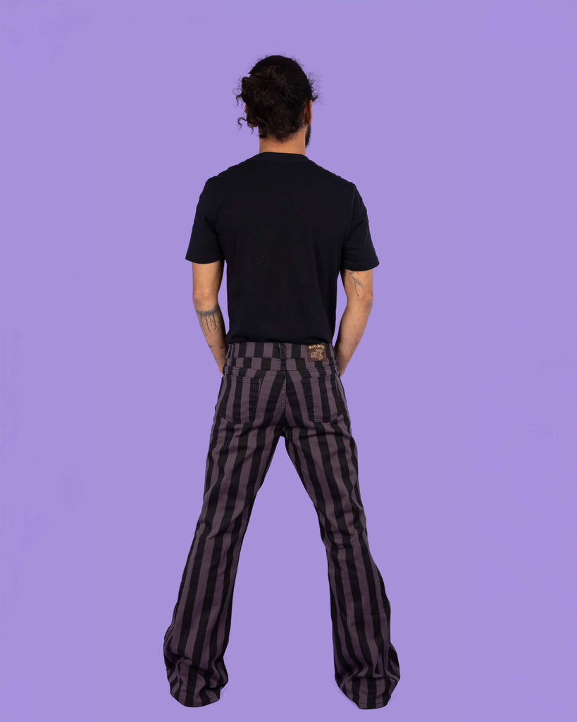 Back view of a model wearing Run & Fly grey and black vertical striped flare trousers. The trousers feature a classic five-pocket design, sitting high on the waist with a comfortable fit and flared legs, ideal for casual wear. Model pairs them with a simple black t-shirt, standing in front of a lilac background. Ethical slow fashion brand. Perfect for alternative styles. Made in England.