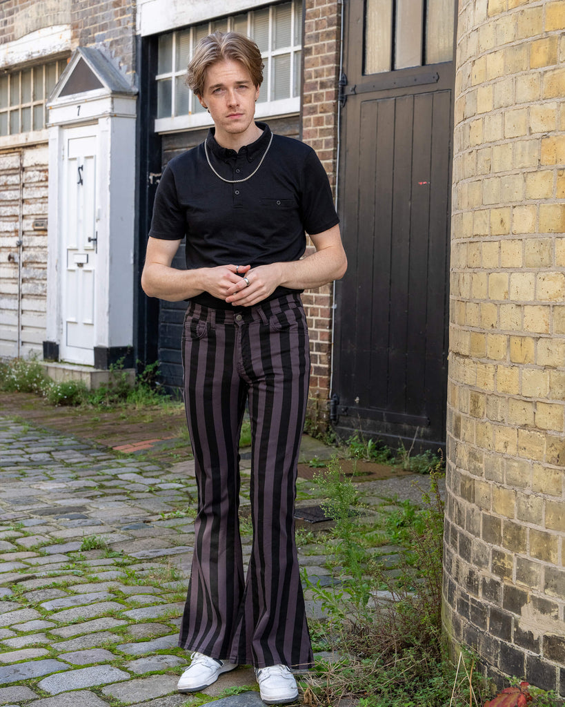 Front view of a model wearing Run & Fly grey and black vertical striped flare trousers. These high-waisted trousers feature a bold flare and vertical stripes for a retro-inspired look, paired with a simple black t-shirt and white trainers. The model stands in a cobblestone alleyway with dark wooden doors. Made ethically by a small slow fashion company in England.