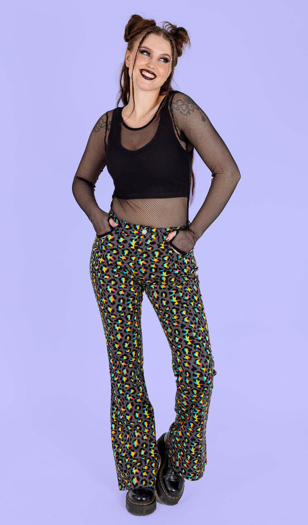 Chloe a femme model with gothic makeup is wearing Run & Fly Grey Rainbow Leopard Print Bell Bottom Flares paired with a black fishnet top and chunky black boots. The flares are a dark grey with an all over rainbow leopard print. Chloe is smiling with her hands in the pockets of the flares and one leg bent in front of a purple background.