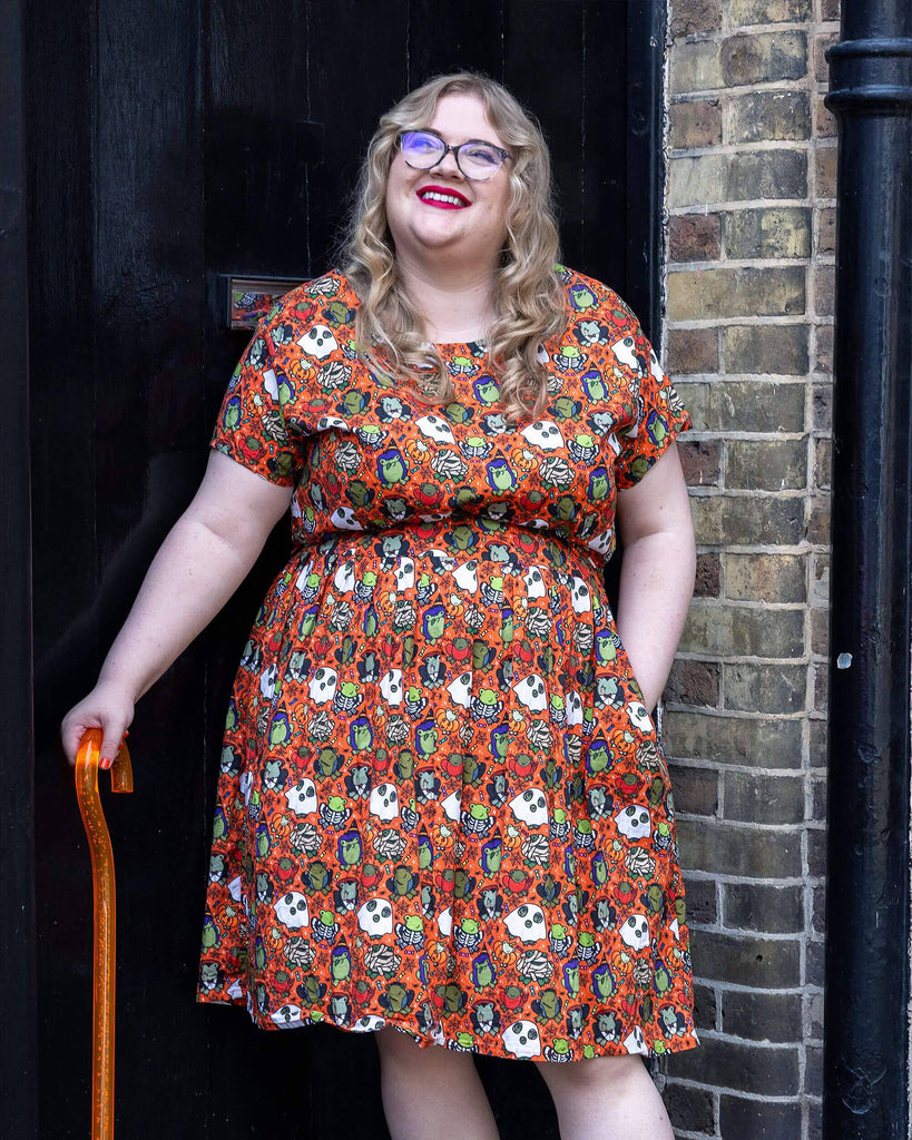 a white femme model with red lipstick wearing Halloween: Party Frogs Stretch Belted Tea Dress with Pockets. The orange dress has an all over print of cartoon frogs in various Halloween costumes such as ghosts and zombies. The model is stood outside in Hove smiling towards the camera with one hand on their hip and other holding an orange walking aid.
