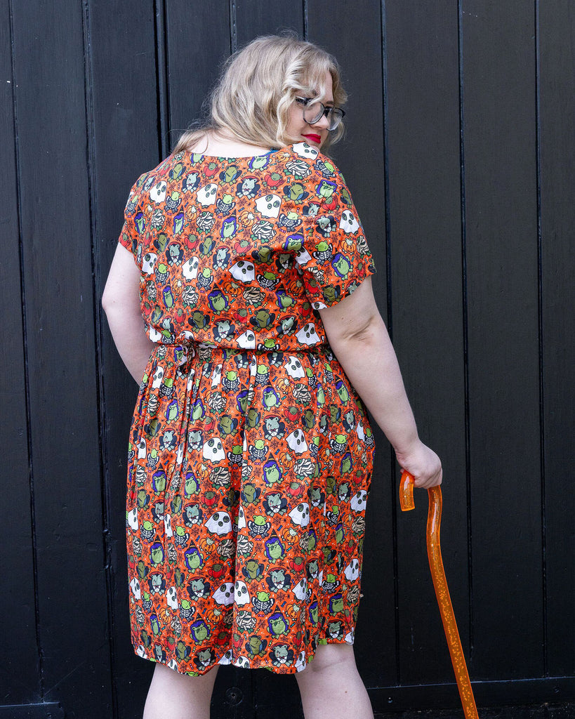 a white femme model with red lipstick wearing Halloween: Party Frogs Stretch Belted Tea Dress with Pockets. The orange dress has an all over print of cartoon frogs in various Halloween costumes such as ghosts and zombies. The model is stood outside in Hove with their back toward the camera to show the back of the dress and is holding an orange walking aid.
