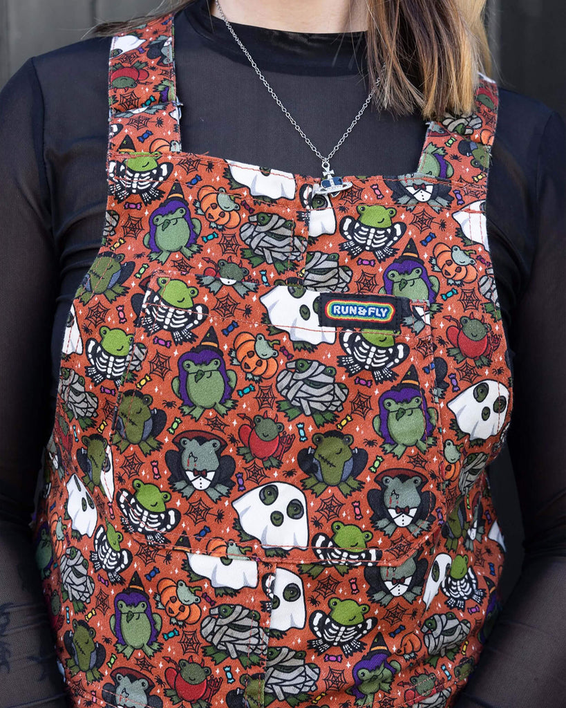 Close up of white female model with blonde hair stood outside in Hove wearing Halloween: Frog Party Stretch Twill Dungarees with a long sleeved black mesh shirt underneath. The dark orange dungarees has an all over print of cartoon frogs in various Halloween costumes such as zombies, vampires and ghosts. The photo is of the waist up, showing the rainbow Run&Fly logo label on the front bib pocket.