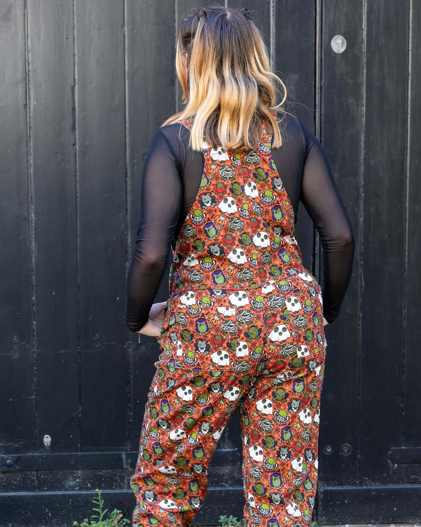 White female model with blonde hair is stood outside in Hove wearing Halloween: Frog Party Stretch Twill Dungarees with a long sleeved black mesh shirt underneath and black boots. The dark orange dungarees has an all over print of cartoon frogs in various Halloween costumes such as zombies, vampires and ghosts. The model is stood with her back to the camera with one leg turned outwards.