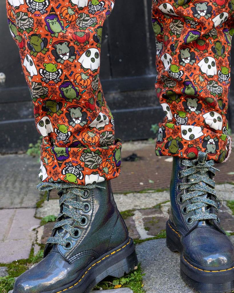 Close up of model stood outside in Hove wearing Halloween: Frog Party Stretch Twill Dungarees with black boots. The dark orange dungarees has an all over print of cartoon frogs in various Halloween costumes such as zombies, vampires and ghosts. Close up shows the matching pattern rolled up legs of the dungarees with the black boots showing underneath. 