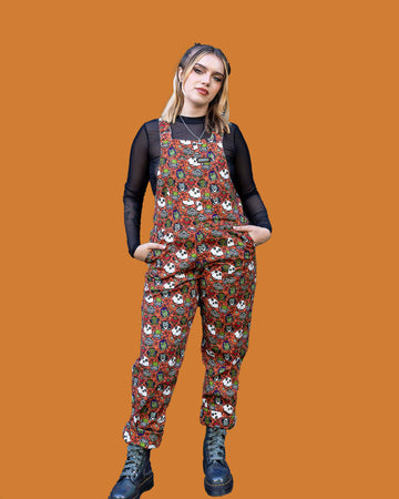 White female model with blonde hair is stood outside in Hove wearing Halloween: Frog Party Stretch Twill Dungarees with a long sleeved black mesh shirt underneath and black boots. The dark orange dungarees has an all over print of cartoon frogs in various Halloween costumes such as zombies, vampires and ghosts. The model is posing toward the camera with her hands in the dungaree front pockets.