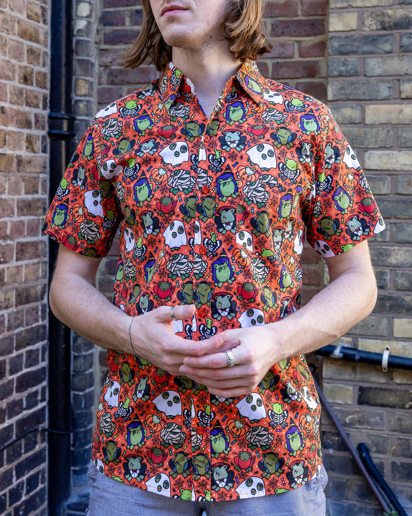 Jack is a tall blonde male model with black round framed glasses wearing an orange shirt with frogs in halloween costumes on it. He is stood in a mews in Hove by a brick wall. He is facing forward adjusting his rings looking off to the left.