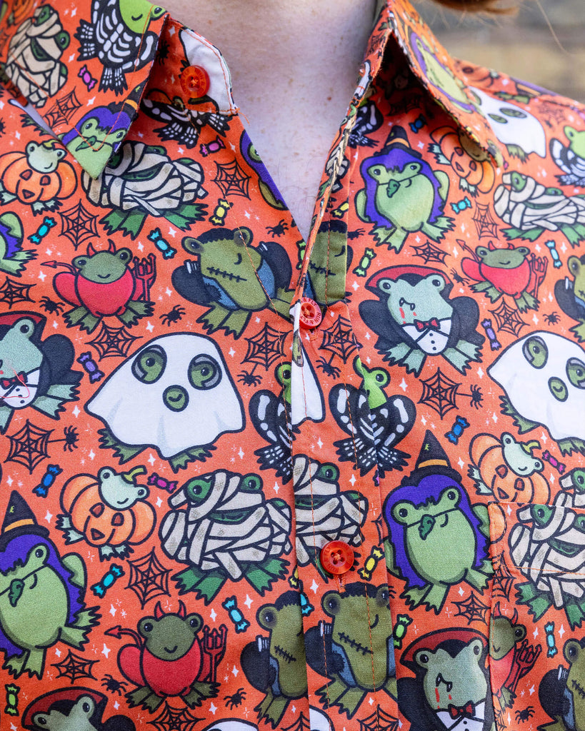 Close up of Jack is a tall blonde male model with black round framed glasses wearing an orange shirt with frogs in halloween costumes on it. He is stood in a mews in Hove by a brick wall. He is facing forward.