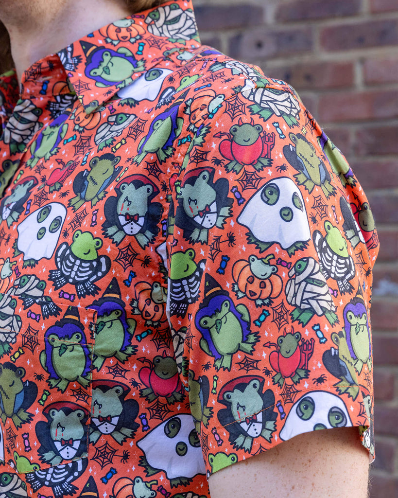 Close up of Jack is a tall blonde male model with black round framed glasses wearing an orange shirt with frogs in halloween costumes on it. He is stood in a mews in Hove by a brick wall. He is facing left to show the shoulder detailing.