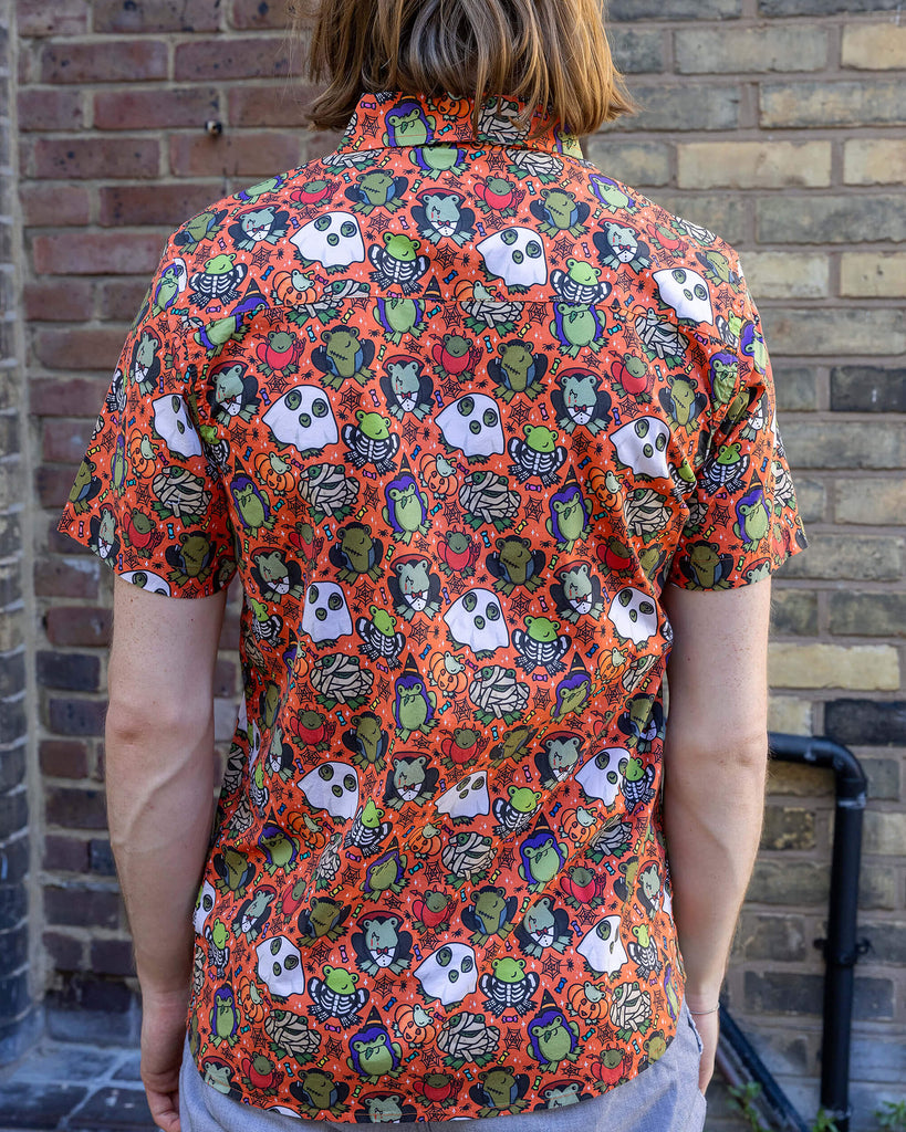 Jack is a tall blonde male model with black round framed glasses wearing an orange shirt with frogs in halloween costumes on it. He is stood in a mews in Hove by a brick wall. He is facing away to show the back of the shirt.