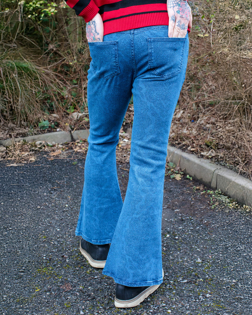 Jim is stood in a garden wearing the retro stretch stone wash denim Hendrix paisley bell bottom flares with a black and red striped jumper and black boat shoes. They are facing away from the camera with one leg forward to highlight the flares with both hands in the back pockets. Photo is cropped from the waist down.