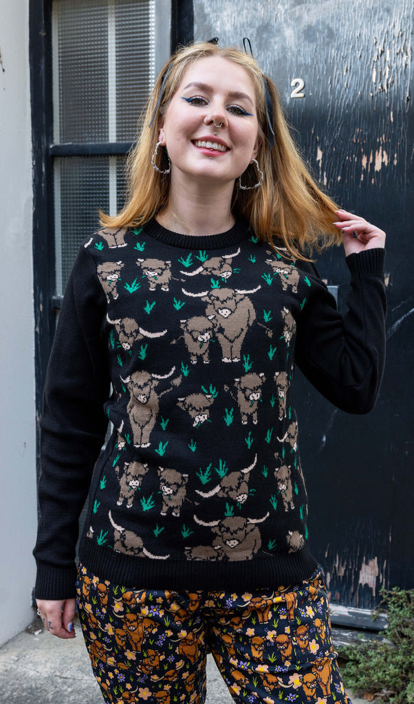 Florence, a femme model with blonde hair is stood outside in Hove wearing Highland Cow Jumper on top of Highland Cow Dungarees. The jumper is black with highland cows on the torso. Florence is smiling facing the camera with one hand in her hair. 