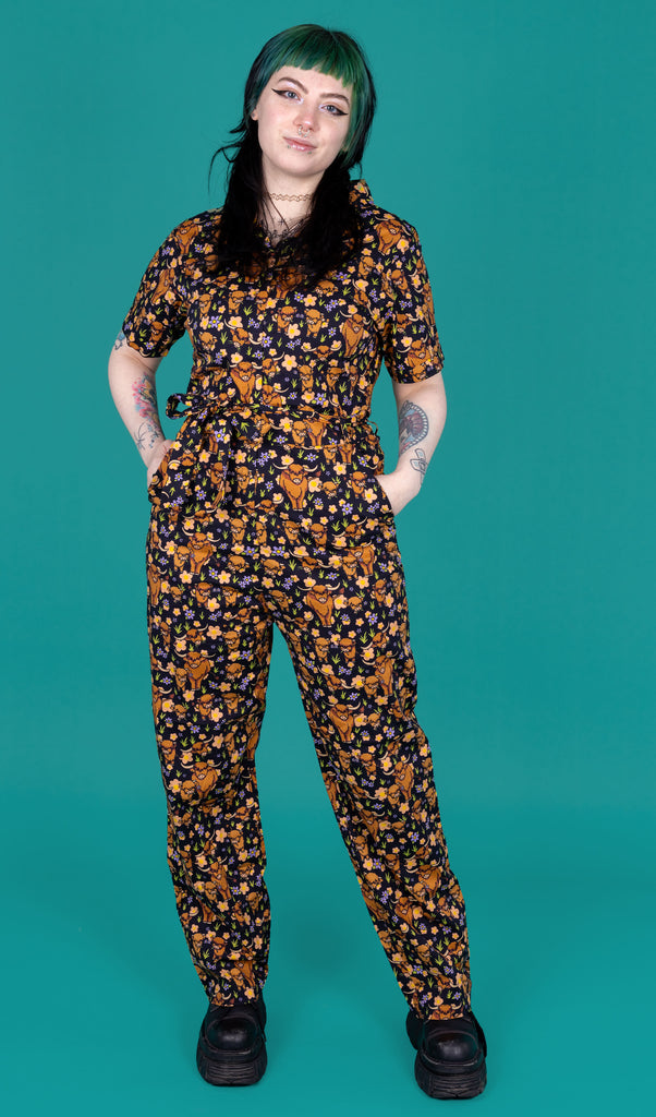 Faeryn, a tattooed model with green and black hair is wearing a black jumpsuit with various sizes of brown highland cows on it, some eating grass. There is also various peach, purple and yellow flowers on the design. The jumpsuit is paired with black boots. The model is posing with their hands in the jumpsuit in front of a green background. 