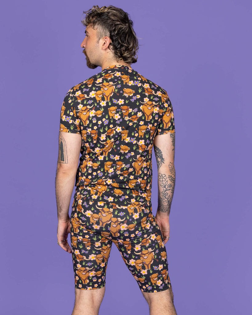 The Highland Cows Stretch Cycle Shorts worn by a masc non binary person with a short brown curly mullet wearing the matching cycle shorts and white trainers on a purple studio background. They are facing away with their arms by their side looking off to the left. The black base shorts features grass, purple and yellow flowers and brown highland cows eating grass.