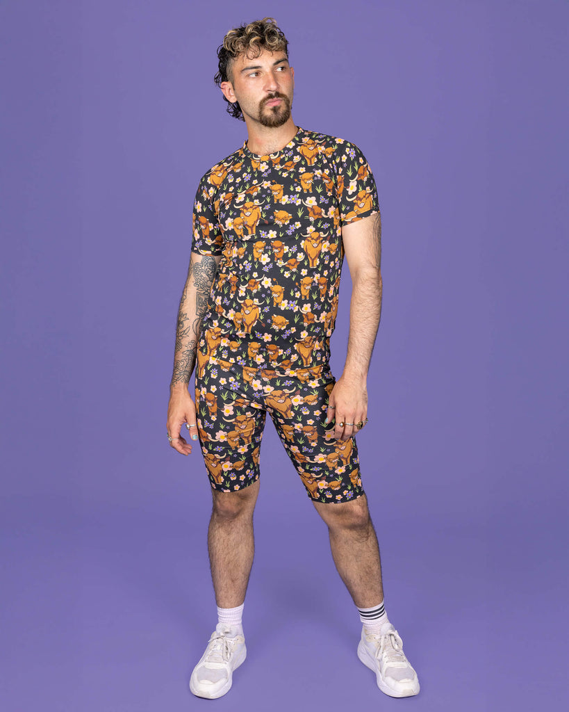 The Highland Cows Stretch Cycle Shorts worn by a masc non binary person with a short brown curly mullet wearing the matching cycle shorts and white trainers on a purple studio background. They are facing forward smising looking off to the right with their arms resting by their side. The black base shorts features grass, purple and yellow flowers and brown highland cows eating grass.