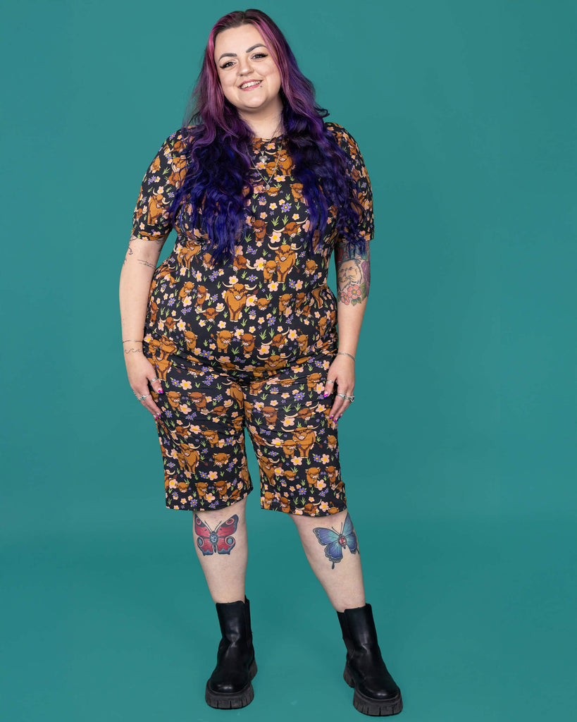 The Highland Cows Stretch Cycle Shorts worn by a femme alternative tattooed model with long pink and purple hair, the matching tshirt and black boots on a teal studio background. She is facing forward smiling with both hands resting by her side. The black base shorts features grass, purple and yellow flowers and brown highland cows eating grass.