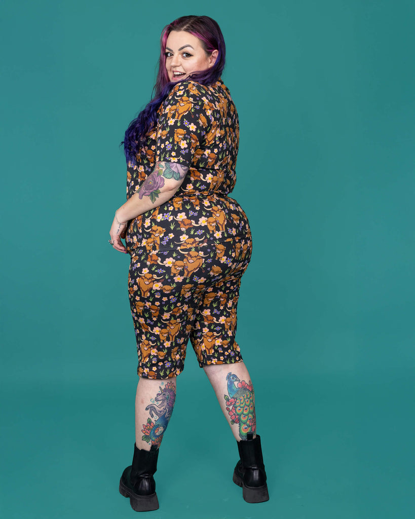 The Highland Cows Stretch Cycle Shorts worn by a femme alternative tattooed model with long pink and purple hair, the matching tshirt and black boots on a teal studio background. She is facing away looking over her left shoulder laughing to camera with both hands resting on her hips. The black base shorts features grass, purple and yellow flowers and brown highland cows eating grass.