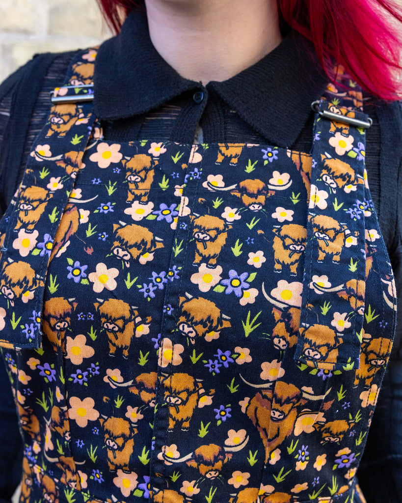 Close up of Florence wearing Highland Cows Stretch Twill Pinafore Dress with a long sleeve black top underneath. The black pinafore dress has an all over print of Highland cows amongst purple flowers and grass designed by Jen James.